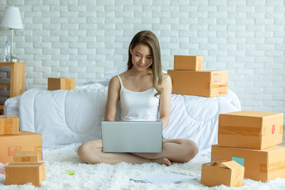 Full length of woman using laptop while sitting amidst cardboard boxes at home