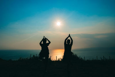 Silhouette friends practicing tree pose at beach against sky during sunset