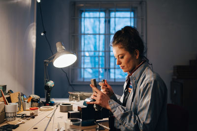 Female engineer working on equipment at illuminated desk by window in workshop