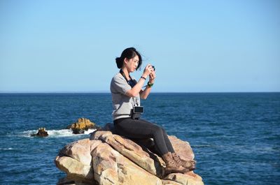 Side view of woman photographing while sitting on rock in sea