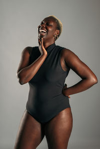 Cheerful african american curvy female in black bodysuit standing on gray background in studio and laughing