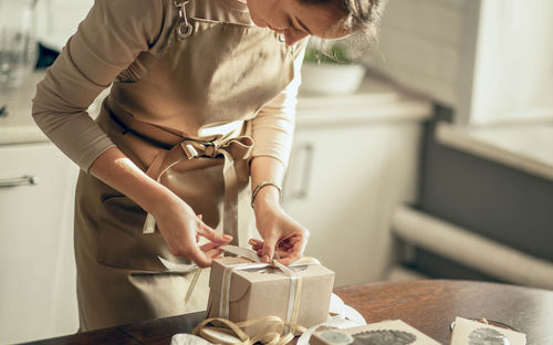 Woman bakery shop owner ties ribbon on box for customer order. chef baking pastry, cake in kitchen