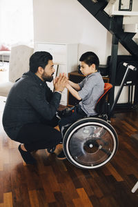 Side view of father holding autistic son's hands sitting on wheelchair at home