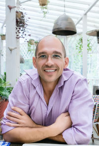 Cheerful bald man with glasses is friendly smiling at the camera, sitting on a summer terrace.