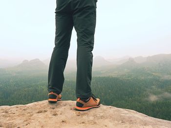 Male legs in dark hiking trousers and leather trekking shoes on peak of rock above misty valley