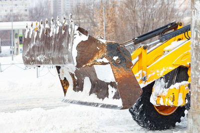 A large metal bucket of a yellow tractor shovels snow from a city road against of a city street.