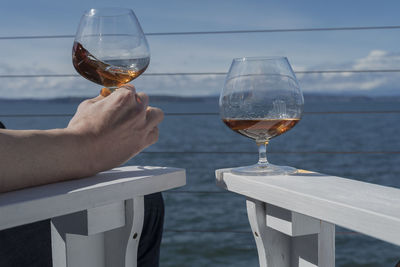 Cropped hand of man holding brandy snifter on white adirondack chairs against sea
