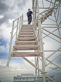 Low angle view of man photographing on smart phone while standing on staircase against sky