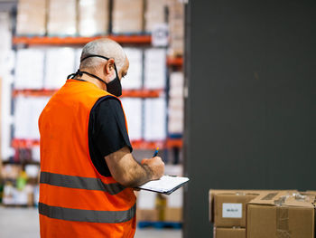 Back view adult worker wearing uniform and protective face mask writing on clipboard while working in spacious storehouse