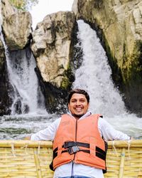 Portrait of smiling young man sitting on wooden raft by rock against waterfall