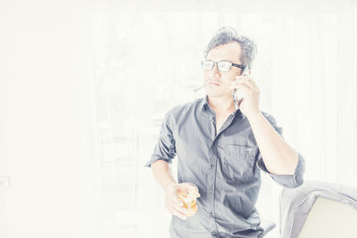 Businessman using smart phone while holding whiskey glass and smoking cigarette in office