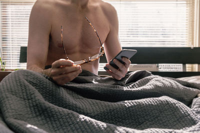 Midsection of man using mobile phone while holding eyeglasses on bed at home