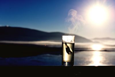 Teabag in glass on railing by lake against bright sky