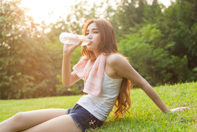 Portrait of woman drinking water from bottle while sitting on field against trees