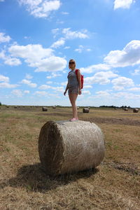 Full length of woman standing on hay bale at agricultural field against sky