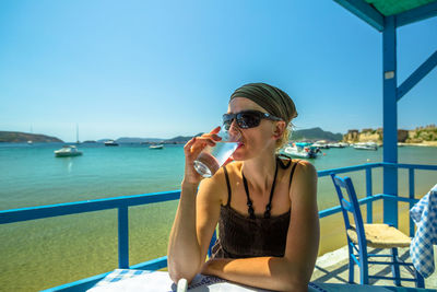 Woman drinking water while sitting at restaurant over sea against clear blue sky