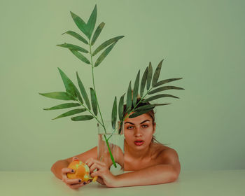 Portrait of young woman with plant and orange fruit against green background