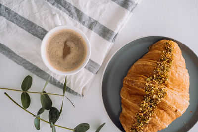 Flat lay of cup of cappuccino with kitchen towel, croissant and eucalyptus branch on the table	
