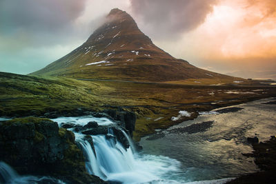 Idyllic view of waterfall and mountain against cloudy sky during sunset