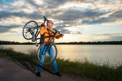 Man standing by bicycle at lakeshore against sky during sunset