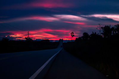 Road amidst silhouette trees against sky at sunset