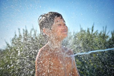 Water spraying on happy shirtless boy against clear blue sky