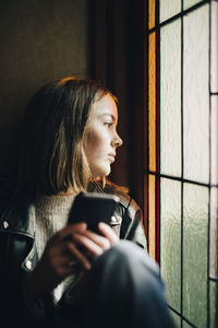 Thoughtful girl with smart phone looking through window while sitting in house