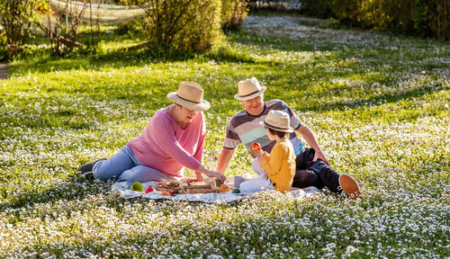 Happy senior family in straw hats having picnic with grandson sitting on blanket on blooming meadow