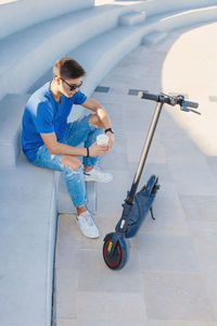 Young caucasian man sitting outdoor near electric scooter and drinking coffee to-go