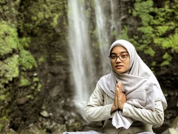 Portrait of young woman with hands clasped sitting against waterfall
