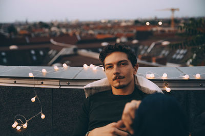 Portrait of young man sitting on illuminated terrace in city during party