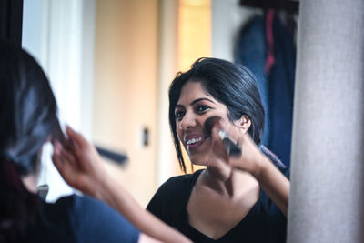 Reflection of smiling woman applying blusher on face in mirror at home