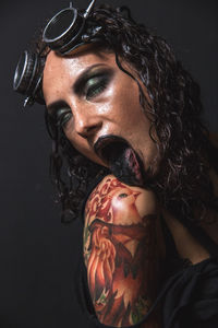 Close-up of woman with make-up licking shoulder with tattoo against black background