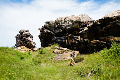 Female hiker running on grassy field by rock formation against cloudy sky during sunny day