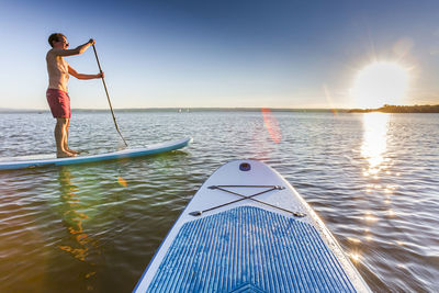 Side view of shirtless man paddleboarding on river against sky during sunset