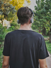 Rear view of man listening to music while standing against trees in park