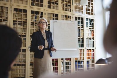 Senior woman giving presentation while standing against bookshelf