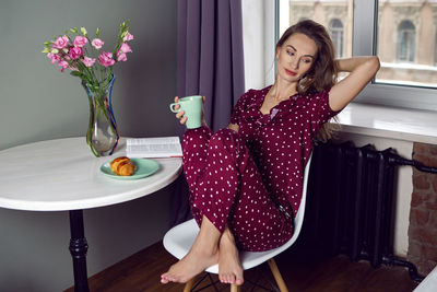 Young woman in red pajamas with polka dots sitting at breakfast at the table next to the window