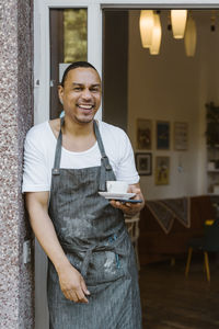 Portrait of happy male owner wearing apron holding coffee cup while leaning at cafe doorway