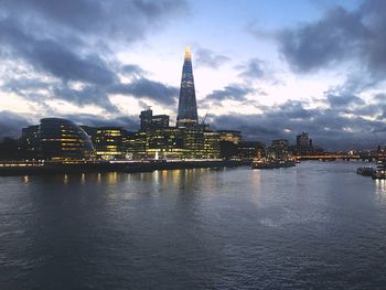 Illuminated buildings by river against cloudy sky