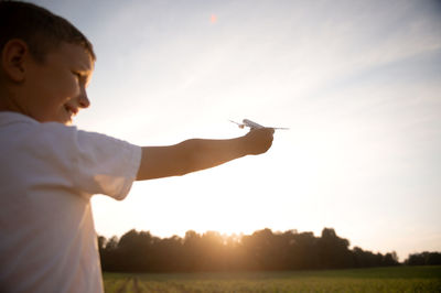 Side view of young man with arms raised standing against sky during sunset