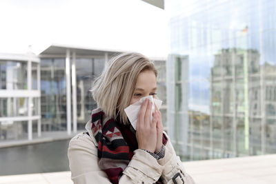 Young woman blowing nose while standing against building 