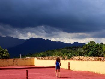Rear view of girl running on building terrace against cloudy sky