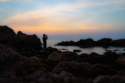 Man standing on rock by sea against sky during sunset