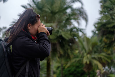 Side view of woman photographing from camera against trees