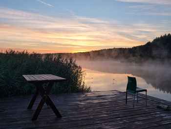 Early morning, lake, fog, beautiful sky. there is an empty table and chair on the wooden dock.