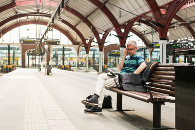 Full length of senior man using technologies while sitting on bench at railroad station