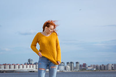 Red-haired woman poses against river in city with buildings in background.