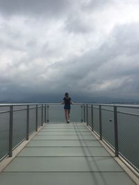 Rear view of woman walking on footbridge over sea against sky