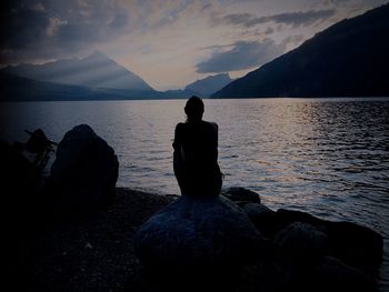 Rear view of silhouette woman sitting on rock by lake against sky
